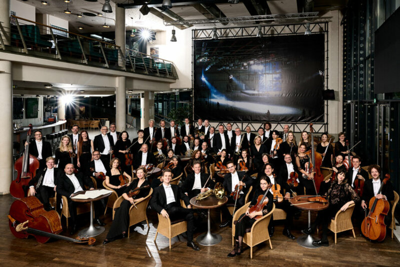 Foto: Torsten Hönig Das gesamte Orchester sitzt zusammen und schaut direkt in die Kamera, die Musiker*innen sind in Konzertkleidung und sind von ihren Instrumenten umgeben.